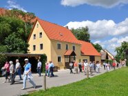 Spazierende Menschen vor einem gelben Haus bei schönem Wetter und Wolken am Himmel. 