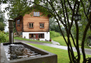 Ein plätschernder Brunnen und ein mehrstämmiger, großer Strauch im Vordergrund rahmen ein Holzhaus ein.